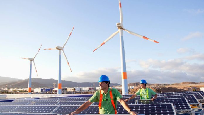 Engineers inspecting solar panels with wind turbines in the background at a renewable energy site.