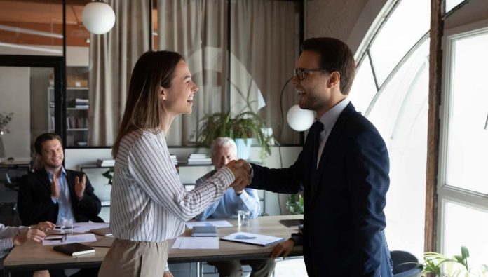 Woman and man shake hands in modern office with colleagues applauding.
