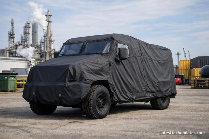 Covered armored vehicle in an industrial setting with refinery backdrop.